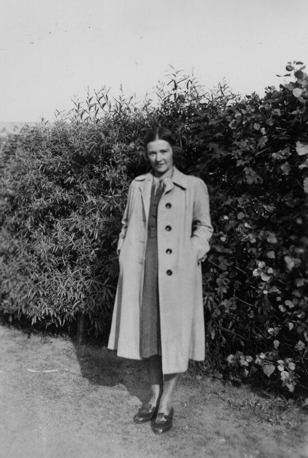 Eine junge Frau mit Mantel steht vor einer Hecke und guckt in die Kamera